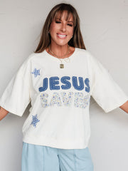 A portrait photograph focusing from the waist up of a smiling woman with long brown hair with straight bangs posing against a textured white wall. She is wearing an oversized, cream-colored short-sleeved sweatshirt made from an 82% cotton and 18% polyester blend fabric, featuring textured patchwork lettering that reads "JESUS SAVES" in blue denim and floral fabrics with blue gingham star patches, styled with light blue seamed pants and layered gold necklaces.