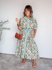 A full-length photograph of a smiling woman with medium-length brown hair and bangs posing with one hand in her dress pocket against a textured white wall. She is wearing a cream-colored midi shirt dress made from 100% cotton fabric featuring a green and blue floral block-print pattern, a geometric-patterned collar, placket, cuffs, and tie belt, paired with brown flat sandals and styled with a brown leather shoulder bag, gold hoop earrings, and stacked gold bangles.