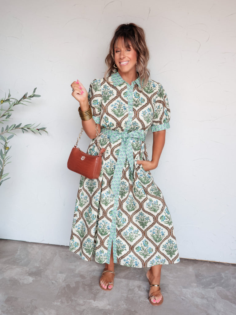 A full-length photograph of a smiling woman with medium-length brown hair and bangs posing with one hand in her dress pocket against a textured white wall. She is wearing a cream-colored midi shirt dress made from 100% cotton fabric featuring a green and blue floral block-print pattern, a geometric-patterned collar, placket, cuffs, and tie belt, paired with brown flat sandals and styled with a brown leather shoulder bag, gold hoop earrings, and stacked gold bangles.