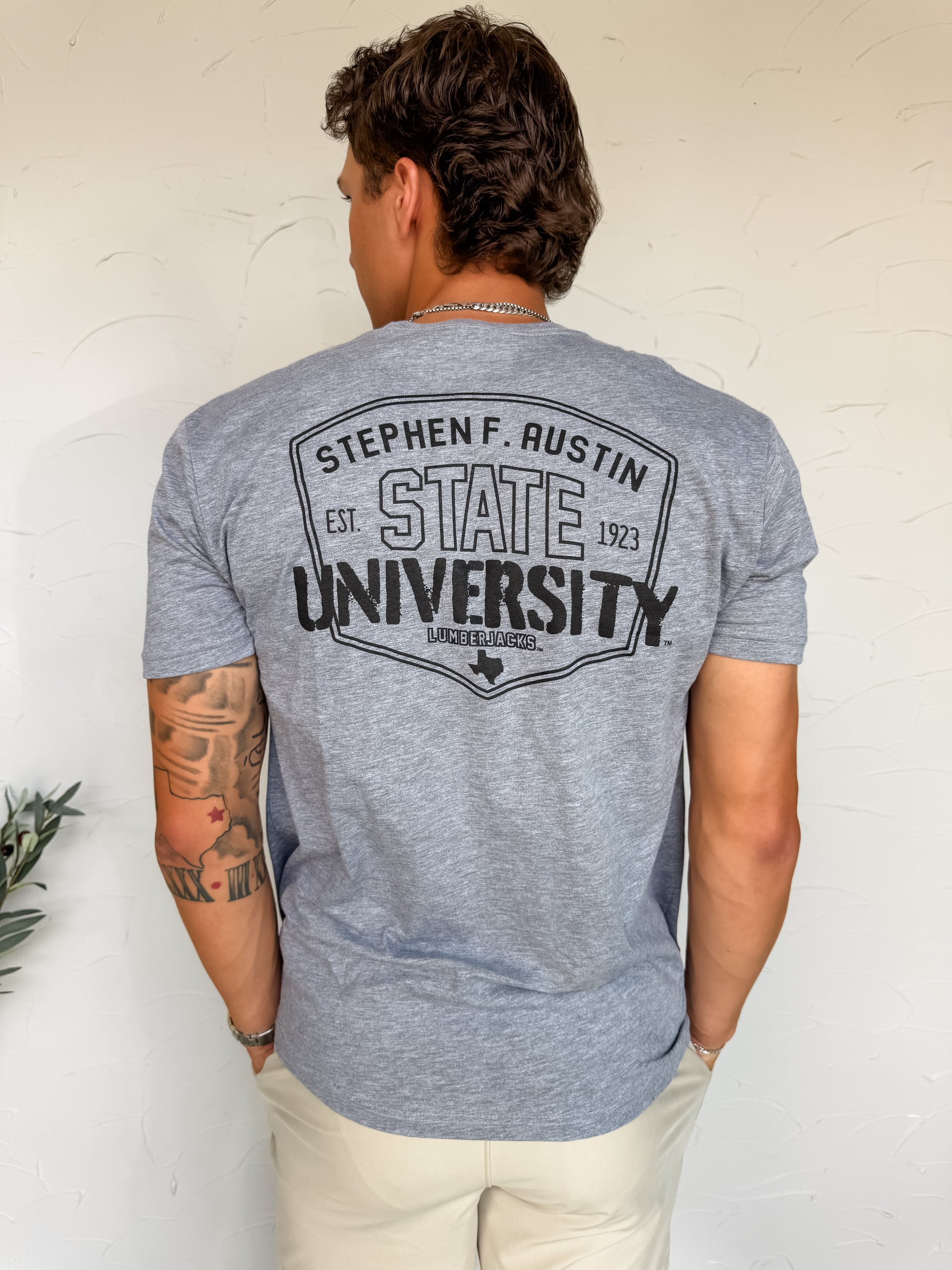 Person wearing a gray t-shirt with 'Stephen F. Austin State University' printed on the back against a white wall.