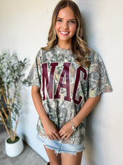 A woman wearing a camouflage graphic t-shirt with 'NAC' printed in maroon letters, paired with denim skirt, standing against a white wall.