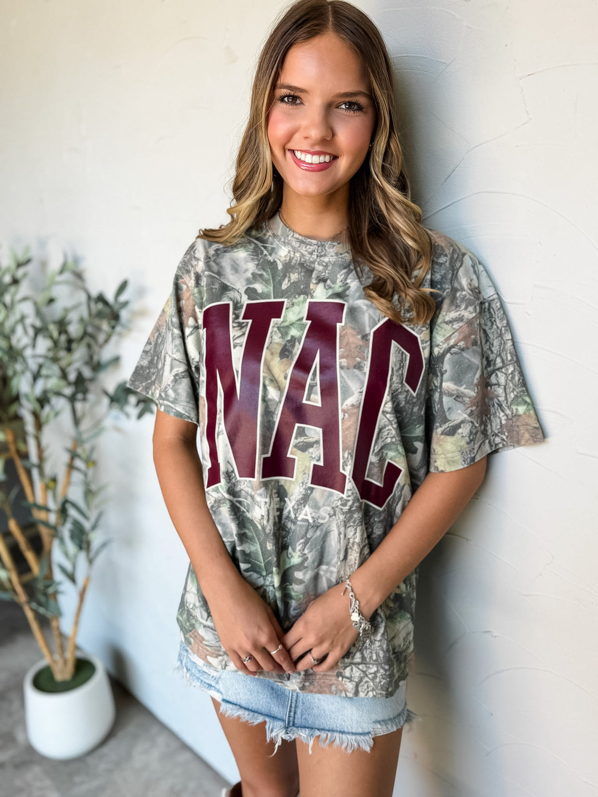 A woman wearing a camouflage graphic t-shirt with 'NAC' printed in maroon letters, paired with denim skirt, standing against a white wall.