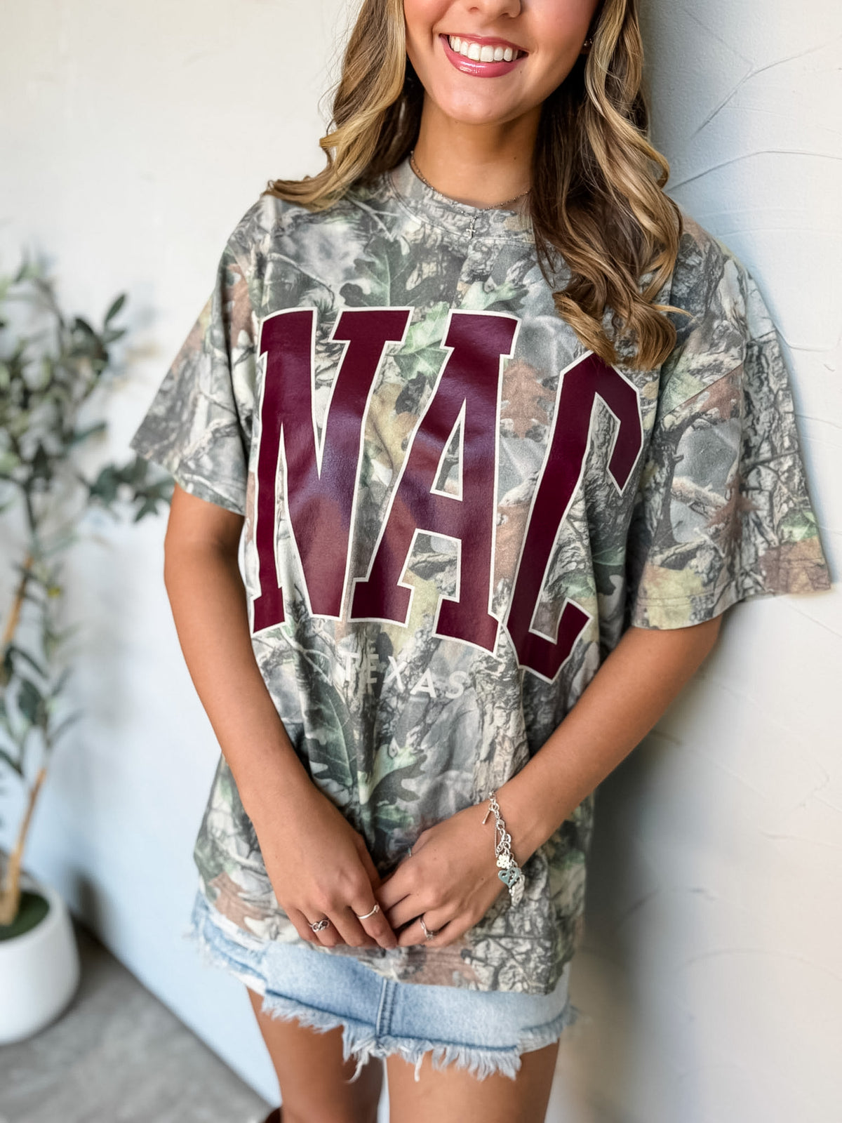A woman wearing a camouflage graphic t-shirt with 'NAC' printed in maroon varsity letters, paired with denim shorts, standing against a white wall.