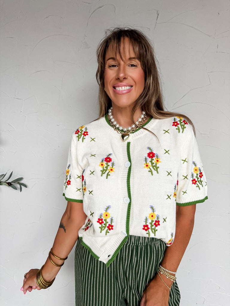 A three-quarter length photograph of a smiling woman with straight brown hair with bangs posing against a textured white wall. She is wearing a short-sleeved cream off-white fine-knit cardigan sweater made from 100% cotton, featuring embroidered red, yellow, and purple wildflower bouquets with scattered green cross-stitch details, contrast green ribbed trim along the crew neck, front button placket, sleeve cuffs, and hem, paired with high-waisted olive green and white pinstriped trousers, styled with a laye