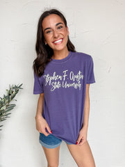 Woman wearing a purple 'Stephen F. Austin State University' t-shirt against a white wall.