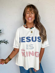 A three-quarter length photograph of a smiling woman with long brown hair and bangs posing against a textured white wall. She is wearing an oversized cream graphic t-shirt made from 100% cotton, featuring patch lettering that reads "JESUS IS KING" with "JESUS" in denim lettering, "IS" in blue floral fabric, and "KING" in pink floral fabric topped with a tiny crown appliqué, paired with blue denim jeans, styled with a colorful beaded necklace, a gold pendant, stacked brass bangles, and beaded bracelets.