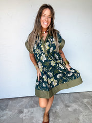 Model wearing the Where The Fern Grows Contrast Trim Floral Dress by Entro in 100% cotton, featuring cream and green botanical florals on a dark blue background, an olive pointed collar and partial button placket, short sleeves with wide olive cuffs, and a relaxed, above-the-knee silhouette with a wide olive hem and contrasting rows of stitching.