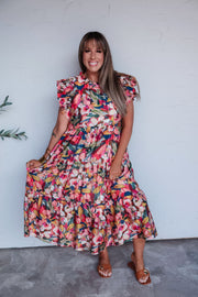 A full-length photograph of a smiling woman with long brown hair and straight bangs posing while holding out the hem of her skirt against a textured white wall. She is wearing a colorful, vibrant floral tiered midi dress made from a 70% rayon and 30% nylon blend fabric, featuring a high round neckline, layered ruffle flutter sleeves with smocked accents, and a relaxed multi-tiered flowy skirt, styled with brown slide sandals, gold bangles, and layered gold necklaces.