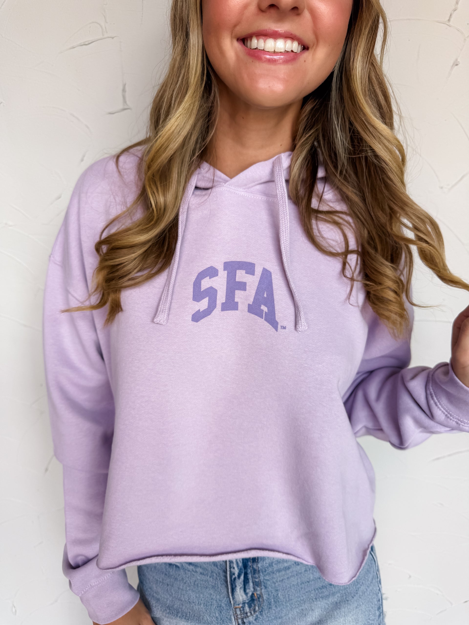 A woman wearing a lavender purple cropped hoodie with 'SFA' printed in a darker purple collegiate font on the chest, paired with blue jeans against a white textured wall.