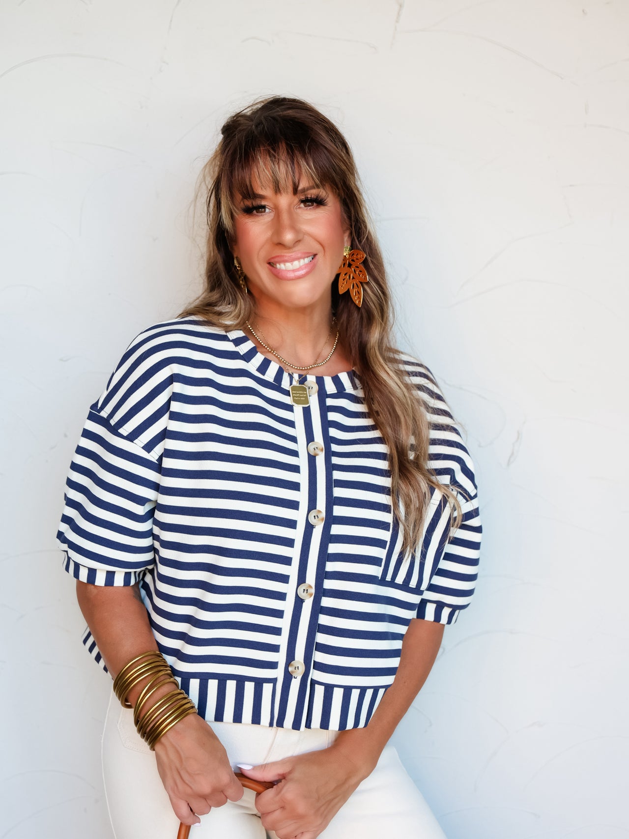 A mid-shot photograph of a smiling brunette woman with bangs posing against a textured white wall. She is wearing a structured navy blue and white horizontally striped short-sleeve cardigan top made from a blend of 62% polyester, 23% cotton, and 15% rayon, featuring a button-down front placket and a chest pocket, styled with white pants and a brown leather handbag.