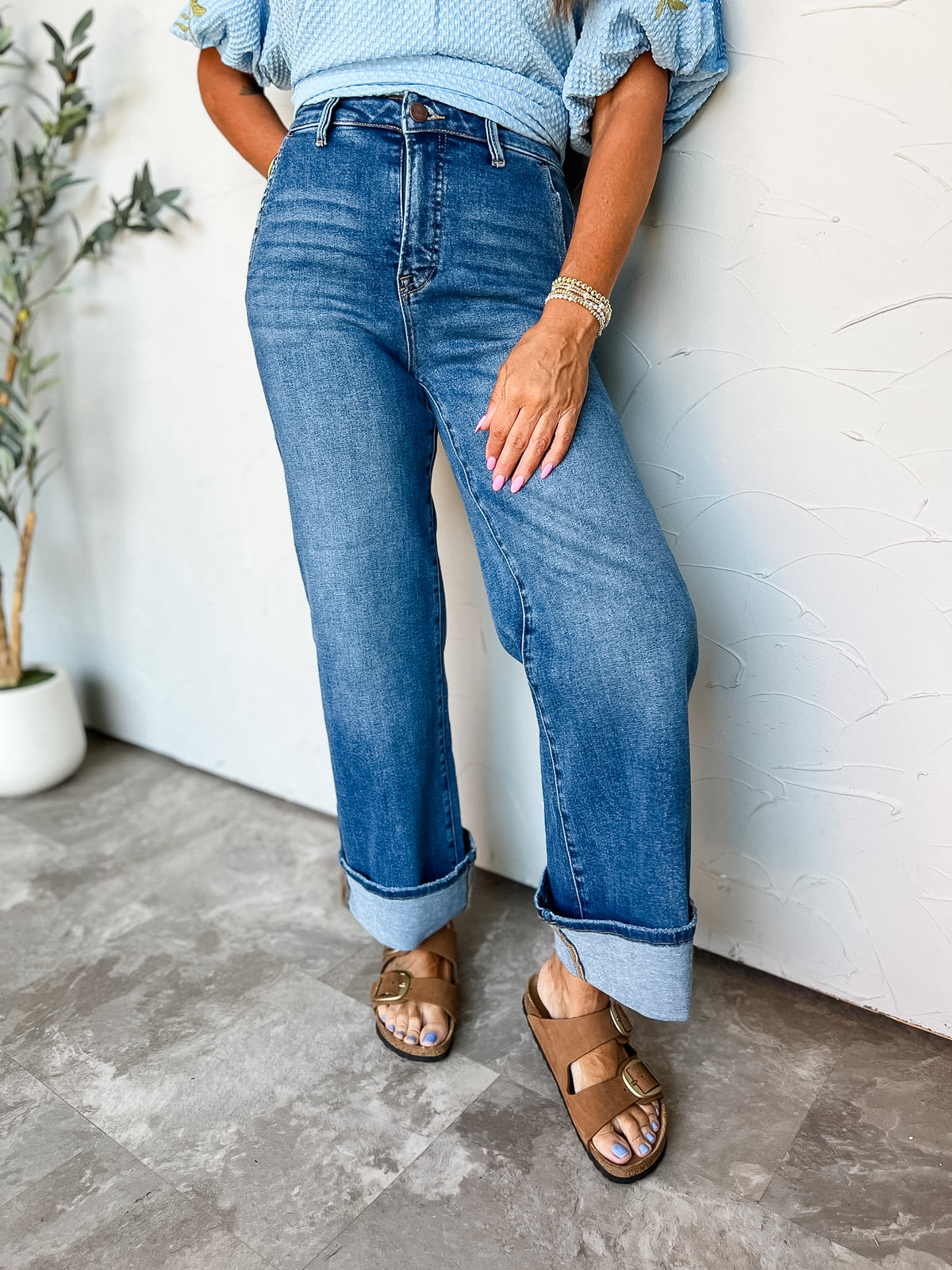 A close-up photograph from the waist down of a person wearing high-rise medium-wash blue denim jeans made from a 93% cotton, 5% polyester, and 2% spandex blend, featuring subtle front whiskering, a relaxed straight-leg fit, and wide folded cuffs at the ankle, paired with brown suede double-buckle slide sandals, tucked under a light blue textured top, styled with beaded gold stretch bracelets.