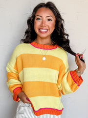 A waist-up photograph of a smiling woman with dark wavy hair touching a strand of her hair while holding the hem of her sweater against a textured white wall. She is wearing a soft knit pullover sweater made from 100% acrylic fabric, featuring wide alternating horizontal stripes in bright yellow and orange with hot pink stitching along the round neckline, ribbed wrist cuffs, and hem, paired with white pinstriped jeans and styled with a round gold pendant necklace and delicate beaded bracelets.