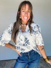 Model wearing the Hold It Together Floral Smocked Ruffle Top by Umgee in 100% polyester, featuring navy floral and paisley motifs on a textured ivory background, a ruffled neckline with a V-shaped opening, smocked detailing, and short puff sleeves with gathered cuffs, styled with dark blue button-front jeans and gold-tone jewelry