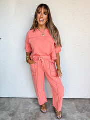A full-length photograph of a smiling woman with straight light-brown hair and front bangs posing with one hand in her pocket against a textured white wall. She is wearing a matching two-piece coral pink lounge set made from an 83% cotton and 17% polyester blend, featuring a relaxed short-sleeved crewneck top with exposed seam detailing, paired with matching straight-leg pants with an elastic drawstring waist and side pockets, styled with brown slide sandals, stacked gold bangles, gold hoop earrings, and a 