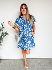 Woman wearing a blue floral dress standing against a white wall.