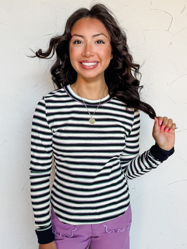 A three-quarter length photograph of a smiling woman with dark wavy hair touching a strand of her hair while posing against a textured white wall. She is wearing a long-sleeved ribbed top made from a 95% polyester and 5% spandex blend, featuring horizontal black, white, purple, and green stripes with solid black cuffs, paired with purple pants with scalloped front pockets and styled with a gold pendant necklace.