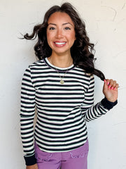 A three-quarter length photograph of a smiling woman with dark wavy hair touching a strand of her hair while posing against a textured white wall. She is wearing a long-sleeved ribbed top made from a 95% polyester and 5% spandex blend, featuring horizontal black, white, purple, and green stripes with solid black cuffs, paired with purple pants with scalloped front pockets and styled with a gold pendant necklace.
