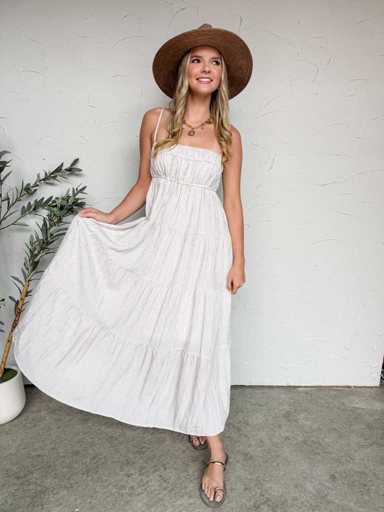 Woman in a white dress and brown hat standing against a white wall with a plant to her left.