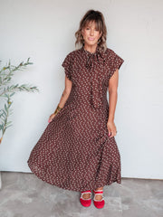 A full-length photograph of a woman with medium-length brown hair and bangs posing against a textured white wall. She is wearing a brown and white polka dot cap-sleeved midi dress made from an 81% viscose and 19% nylon blend, featuring a tied bow at the neckline, paired with red Mary Jane flats with white trim and styled with layered necklaces, gold hoop earrings, and stacked gold bangles.