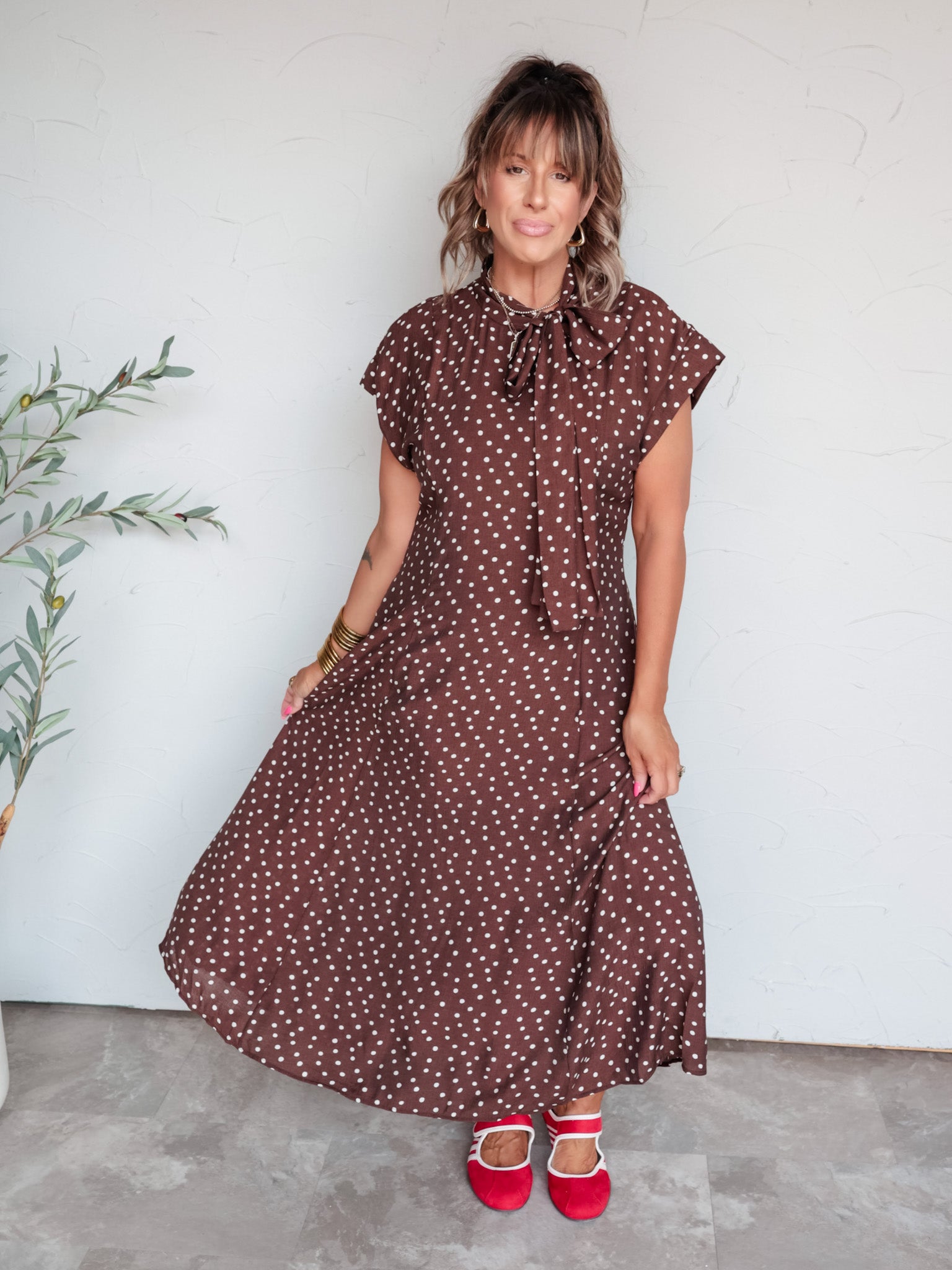A full-length photograph of a woman with medium-length brown hair and bangs posing against a textured white wall. She is wearing a brown and white polka dot cap-sleeved midi dress made from an 81% viscose and 19% nylon blend, featuring a tied bow at the neckline, paired with red Mary Jane flats with white trim and styled with layered necklaces, gold hoop earrings, and stacked gold bangles.