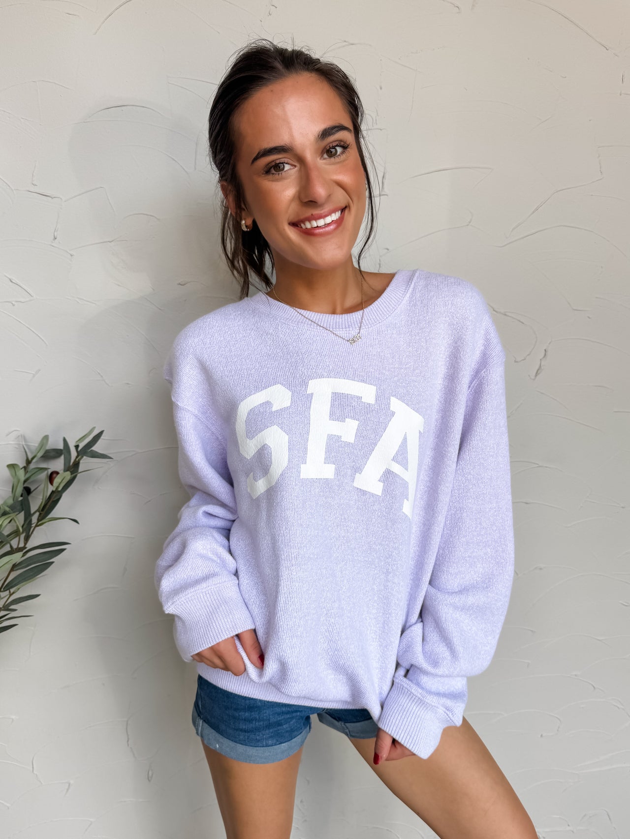 Woman wearing a light purple sweatshirt with 'SFA' on it against a white wall.