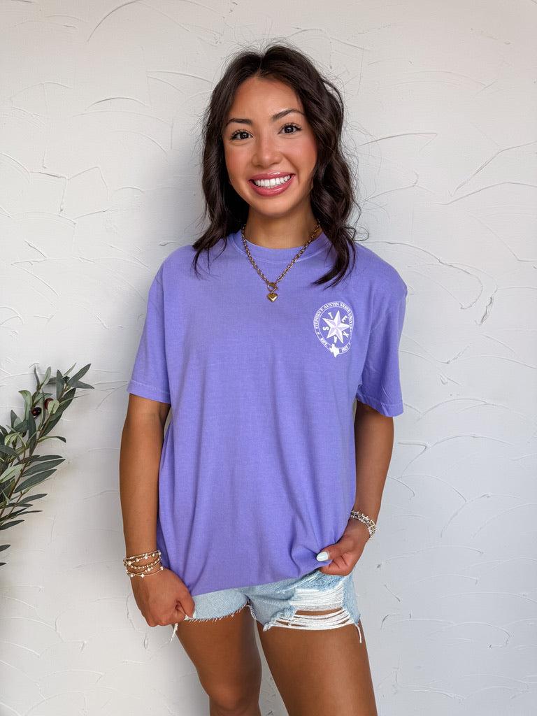 Woman wearing a purple t-shirt with a white logo against a white wall.