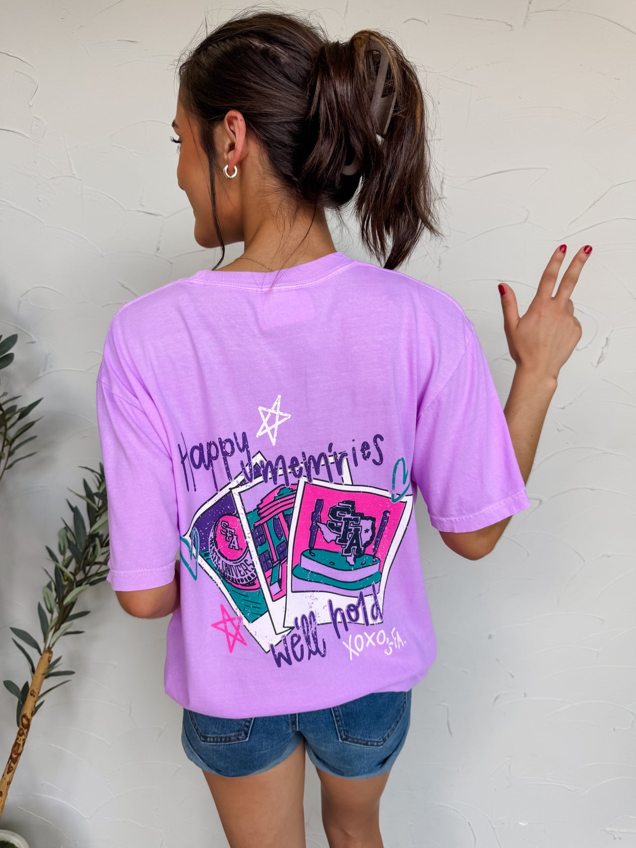 Person wearing a purple t-shirt with graphic designs and text, standing against a white wall.