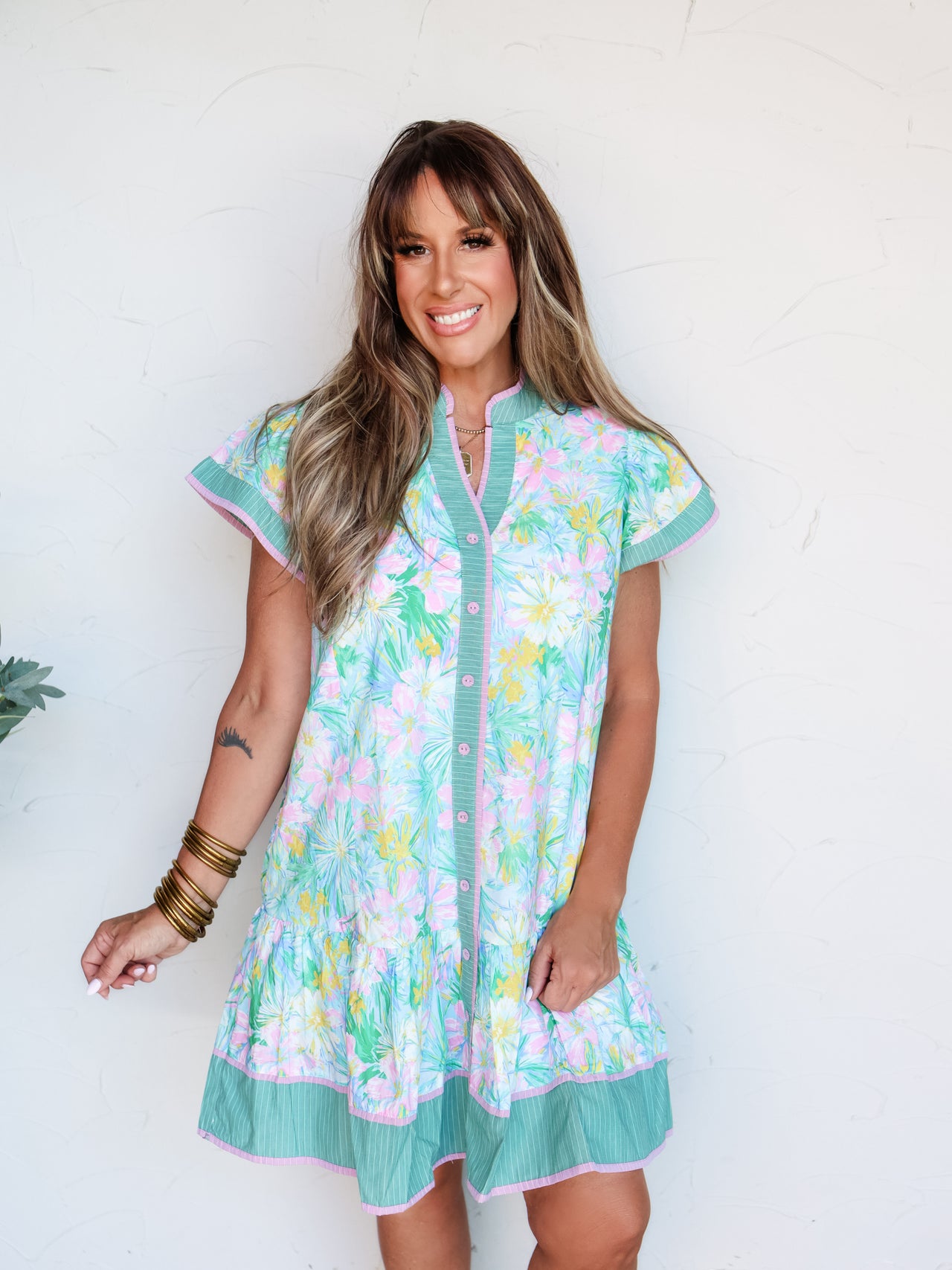 A full-length photograph of a smiling brunette woman with bangs posing with both hands in her pockets against a textured white wall. She is wearing a relaxed multicolor short-sleeve button-down shift dress made from a 100% cotton fabric featuring a turquoise, pink, and yellow floral print, detailed with contrasting green striped trim, pink piping, and a tiered hemline, styled with stacked gold bangles on her wrist.