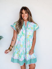 A full-length photograph of a smiling brunette woman with bangs posing with both hands in her pockets against a textured white wall. She is wearing a relaxed multicolor short-sleeve button-down shift dress made from a 100% cotton fabric featuring a turquoise, pink, and yellow floral print, detailed with contrasting green striped trim, pink piping, and a tiered hemline, styled with stacked gold bangles on her wrist.