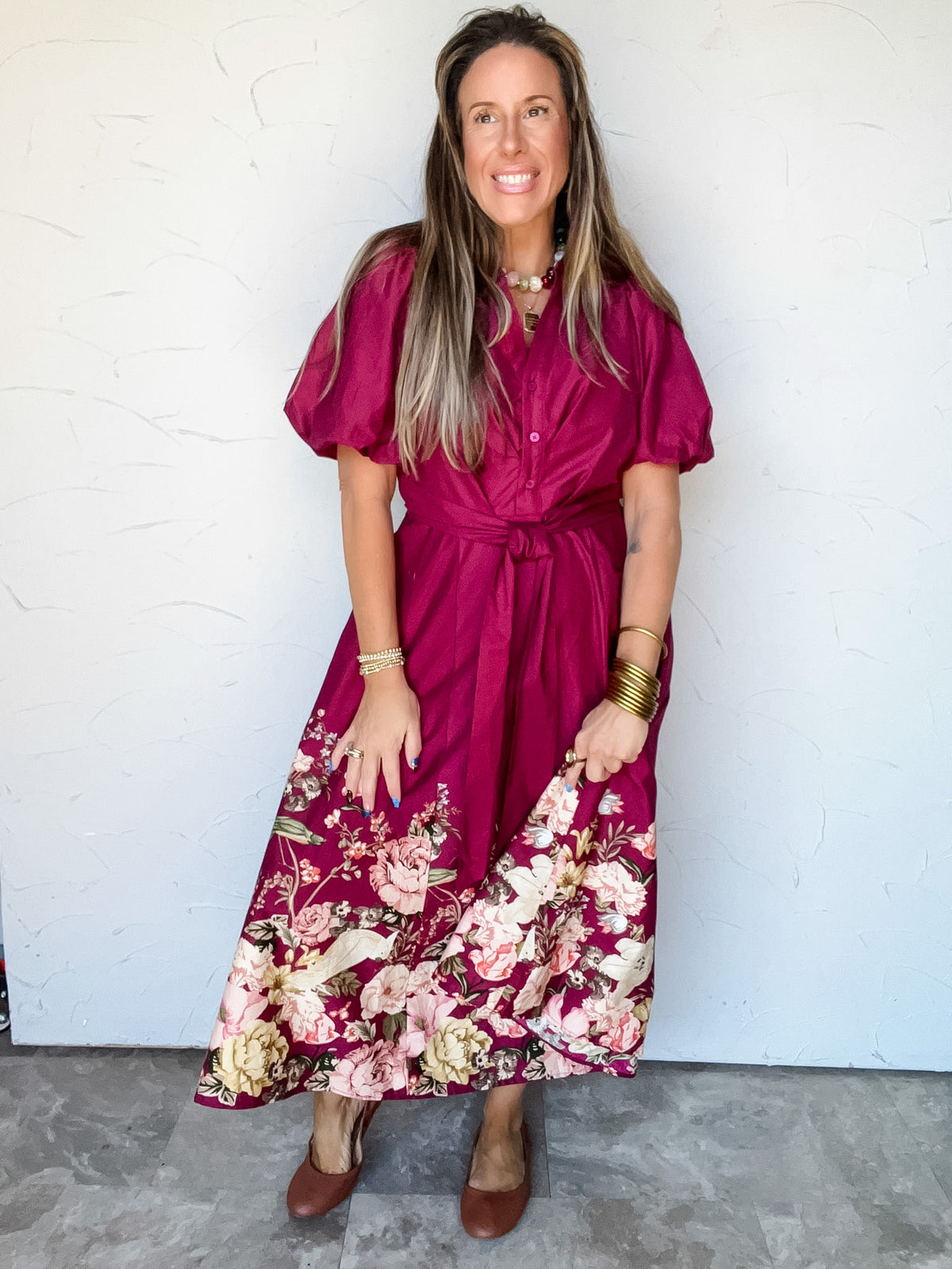 Model wearing the Paint Me A Pretty Picture Floral Border Midi Dress by Entro in 100% cotton, featuring a burgundy button-front bodice, pointed collar, short puff sleeves, bow-tied waist, side pockets, and a flared midi skirt with a cream, blush pink, and yellow floral border.
