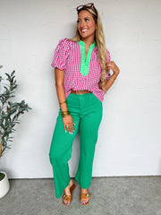 Woman wearing a red and white checkered blouse and green pants against a white wall.