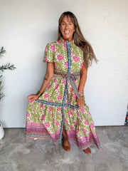 Lesly modeling the t.c.e.c. We Love This One Self Tie Floral Dress in 100% cotton, featuring pink flowers and green leafy accents on a light background, contrasting navy and pink patterned borders, short sleeves, a stand collar, a self-tie waist, and a long tiered skirt with a decorative hem, paired with brown flats.