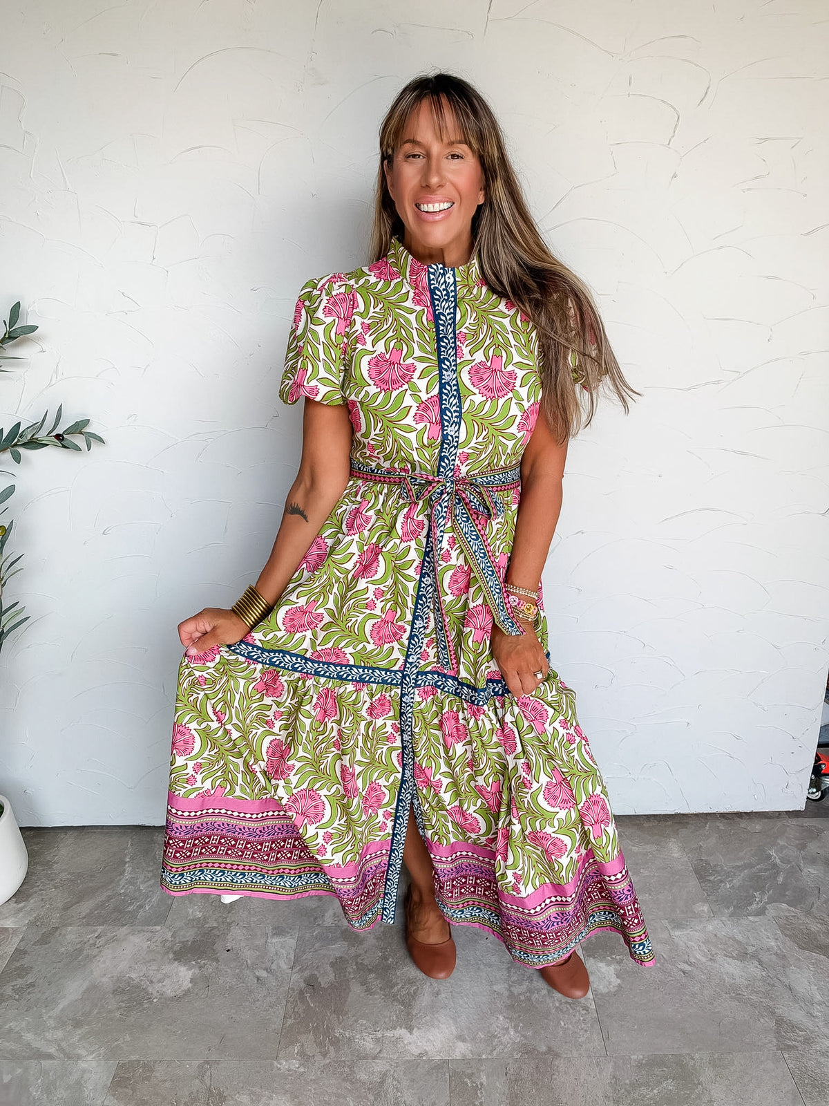 Lesly modeling the t.c.e.c. We Love This One Self Tie Floral Dress in 100% cotton, featuring pink flowers and green leafy accents on a light background, contrasting navy and pink patterned borders, short sleeves, a stand collar, a self-tie waist, and a long tiered skirt with a decorative hem, paired with brown flats.