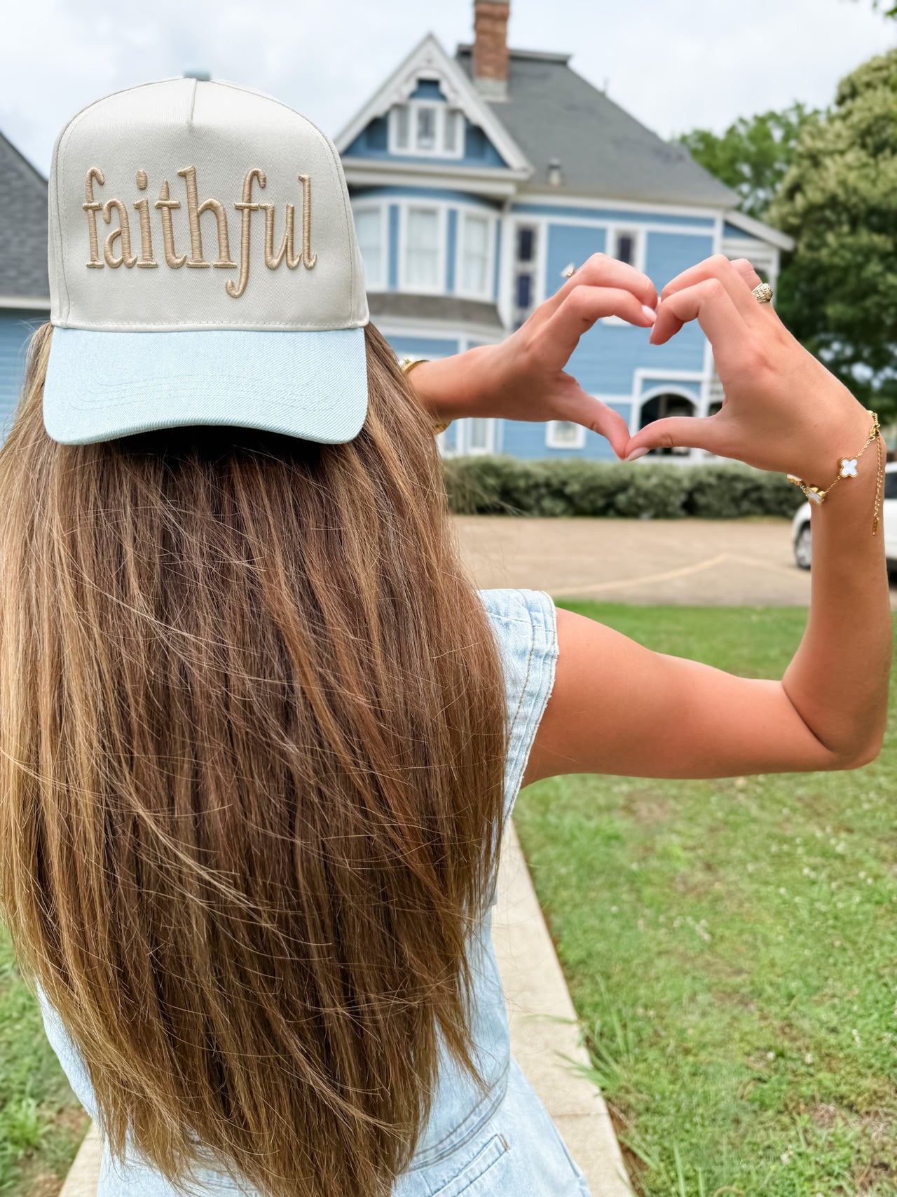 Person wearing a cap with 'faithful' text, making a heart shape with their hands outdoors.