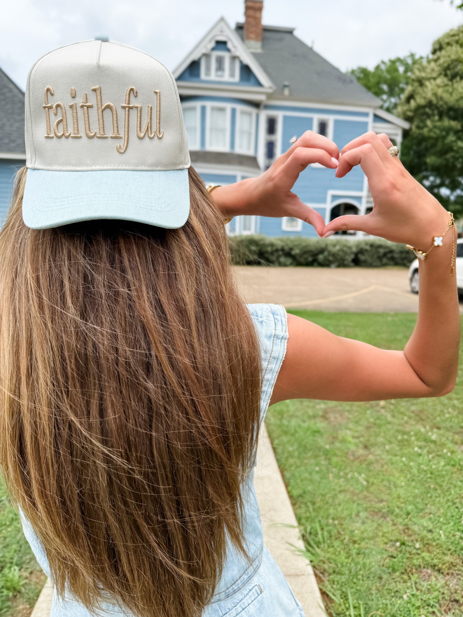 Person wearing a cap with 'faithful' text, making a heart shape with their hands outdoors.