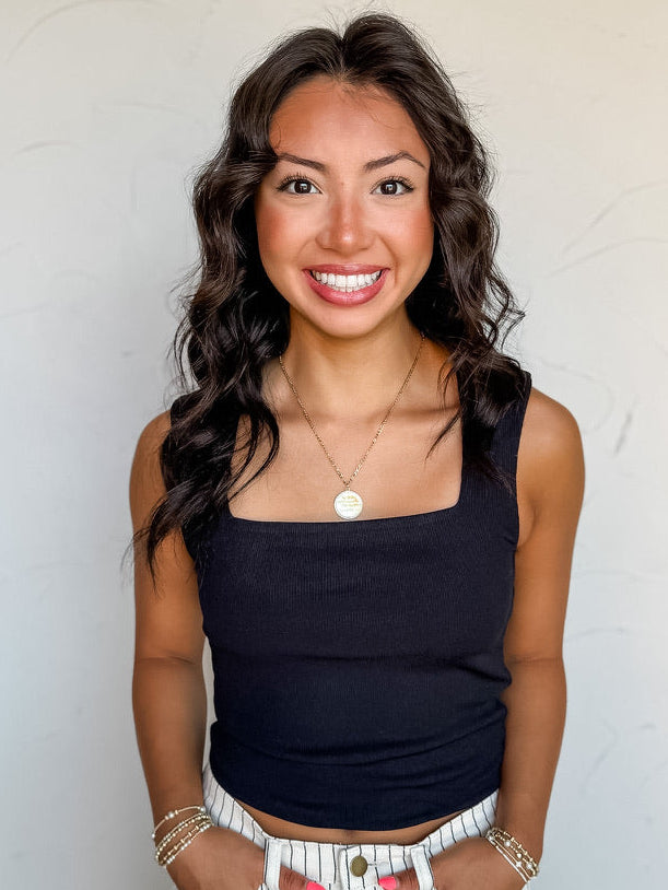 A portrait-style photograph of a smiling woman with dark wavy hair posing with both hands in her pockets against a textured white wall. She is wearing a fitted black ribbed tank top made from a 96% polyester and 4% spandex blend, featuring a square neckline and wide shoulder straps, paired with white pinstriped jeans and styled with a round gold pendant necklace and delicate beaded bracelets.