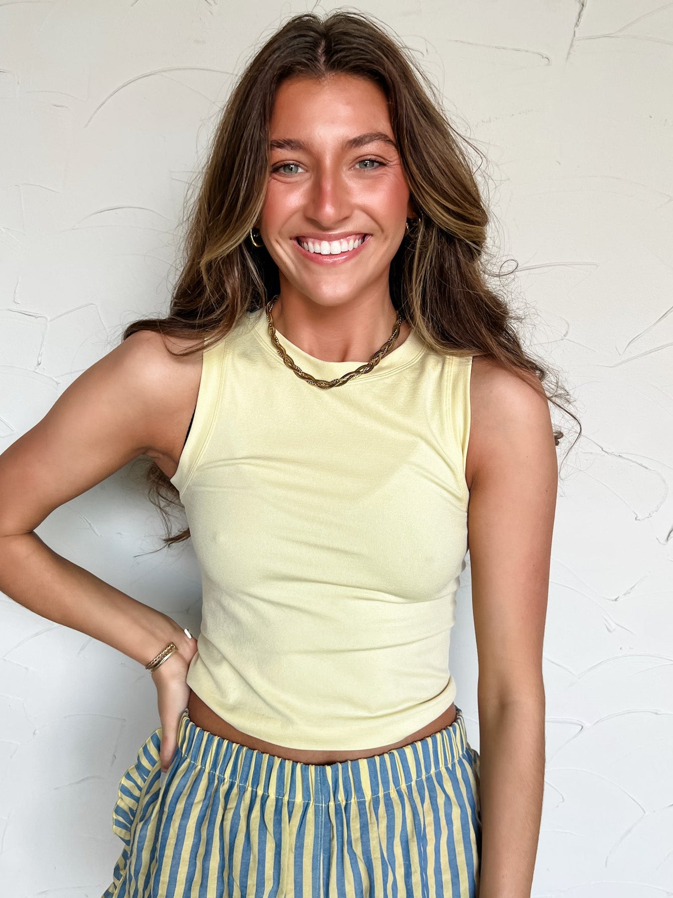 Woman wearing a yellow sleeveless top and striped shorts against a textured white wall.