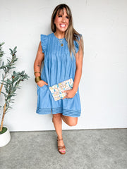 Woman wearing a blue dress holding a floral clutch against a white wall.