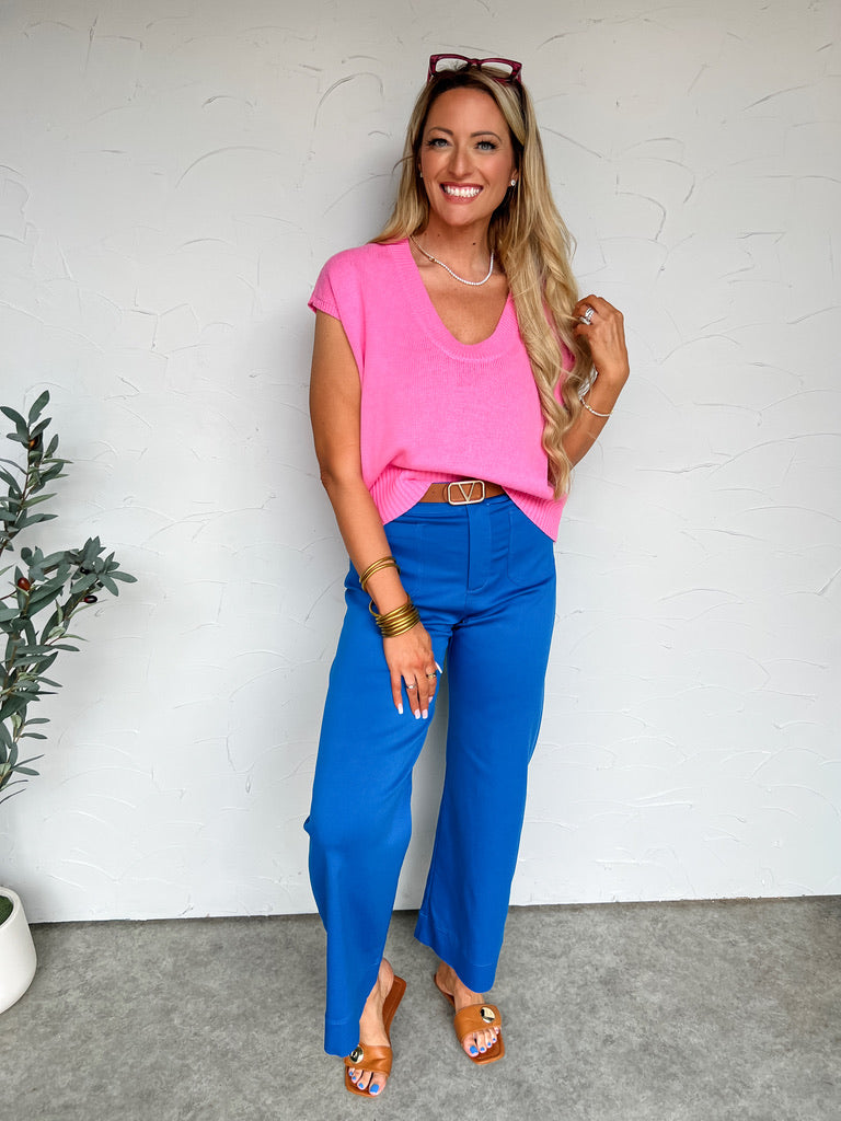 Woman wearing a pink top and blue pants standing against a white wall.