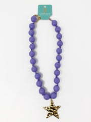 A purple beaded necklace with a gold starfish pendant, hanging from a teal brand tag that reads 'TATUM SHAVE ANIMALS'.