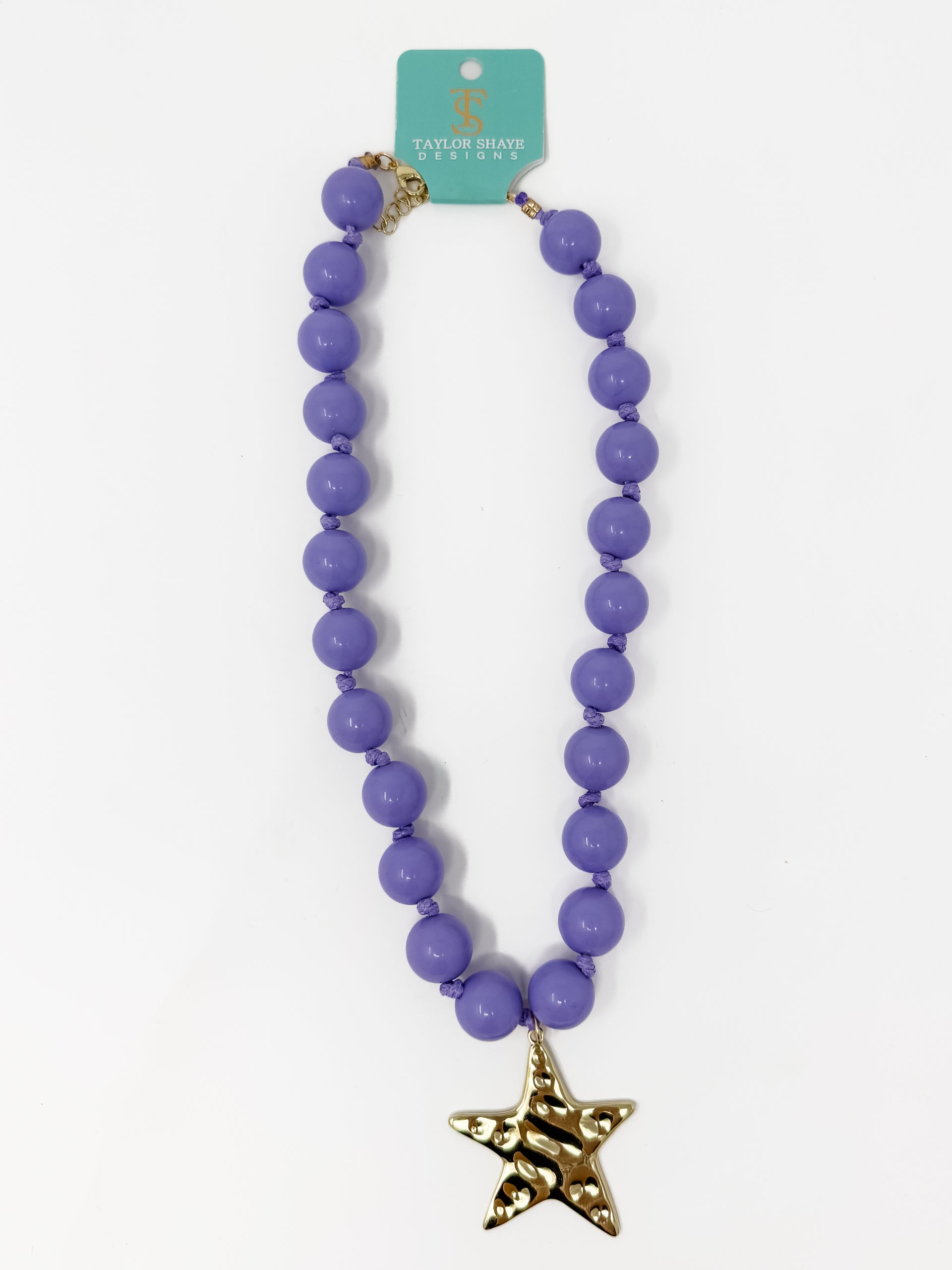 A purple beaded necklace with a gold starfish pendant, hanging from a teal brand tag that reads 'TATUM SHAVE ANIMALS'.