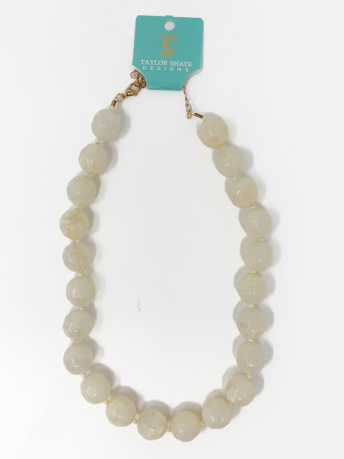 A beaded necklace made of round, polished cream-colored beads with a gold-tone clasp, attached to a teal card that reads 'Taylor Shaye Designs', photographed on a white background.