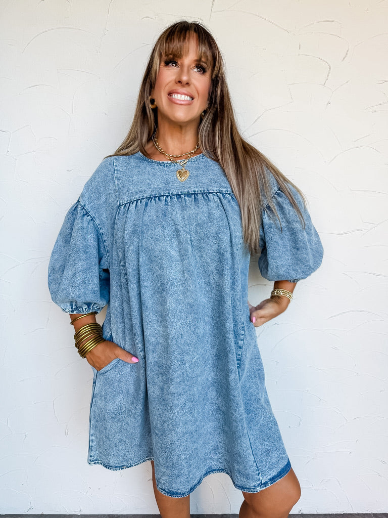 bangs posing with one hand in her pocket against a textured white wall. She is wearing an acid-wash blue denim mini dress made from 100% cotton, featuring a round neckline, gathered empire seam, dramatic elbow-length puff sleeves with elastic cuffs, and side pockets, styled with stacked gold bangles, gold hoop earrings, a gold chain necklace with a heart pendant, and a gold beaded bracelet.