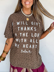 A woman wearing a 100% Cotton brown leopard-print t-shirt with white text that reads 'I WILL GIVE THANKS TO THE LORD WITH ALL MY HEART Psalm 9:1' against a white wall.