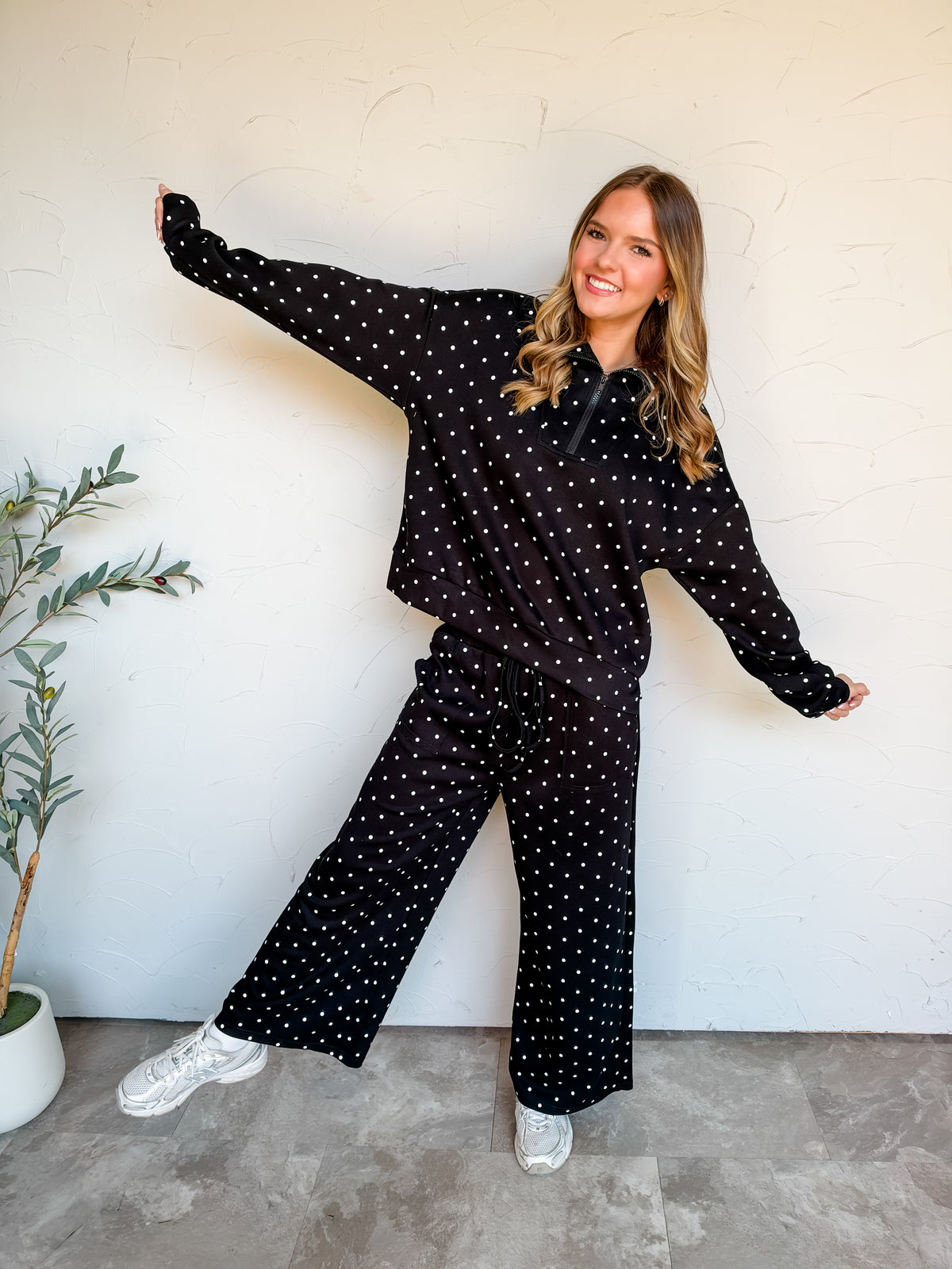 Catie wearing the Calling On You Polka Dot Half Zip Pullover Set in black with small white polka dots, featuring a collared long-sleeve pullover and matching wide-leg pants. Made of 74% polyester, 20% rayon, and 6% spandex, styled with metallic sneakers.⠋

