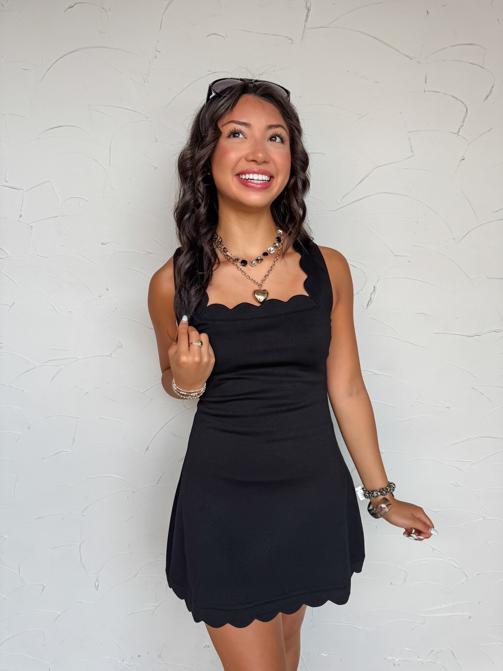 A sleeveless, solid black active dress made from a blend of 49% polyester, 45% rayon, and 6% spandex. The dress is designed with a distinctive scalloped square neckline, wide tank-style shoulder straps, and a fitted bodice that flows into a slight A-line mini skirt. The bottom hem of the skirt features matching scalloped trim detailing, and the dress is fully lined with hidden, built-in shorts underneath.