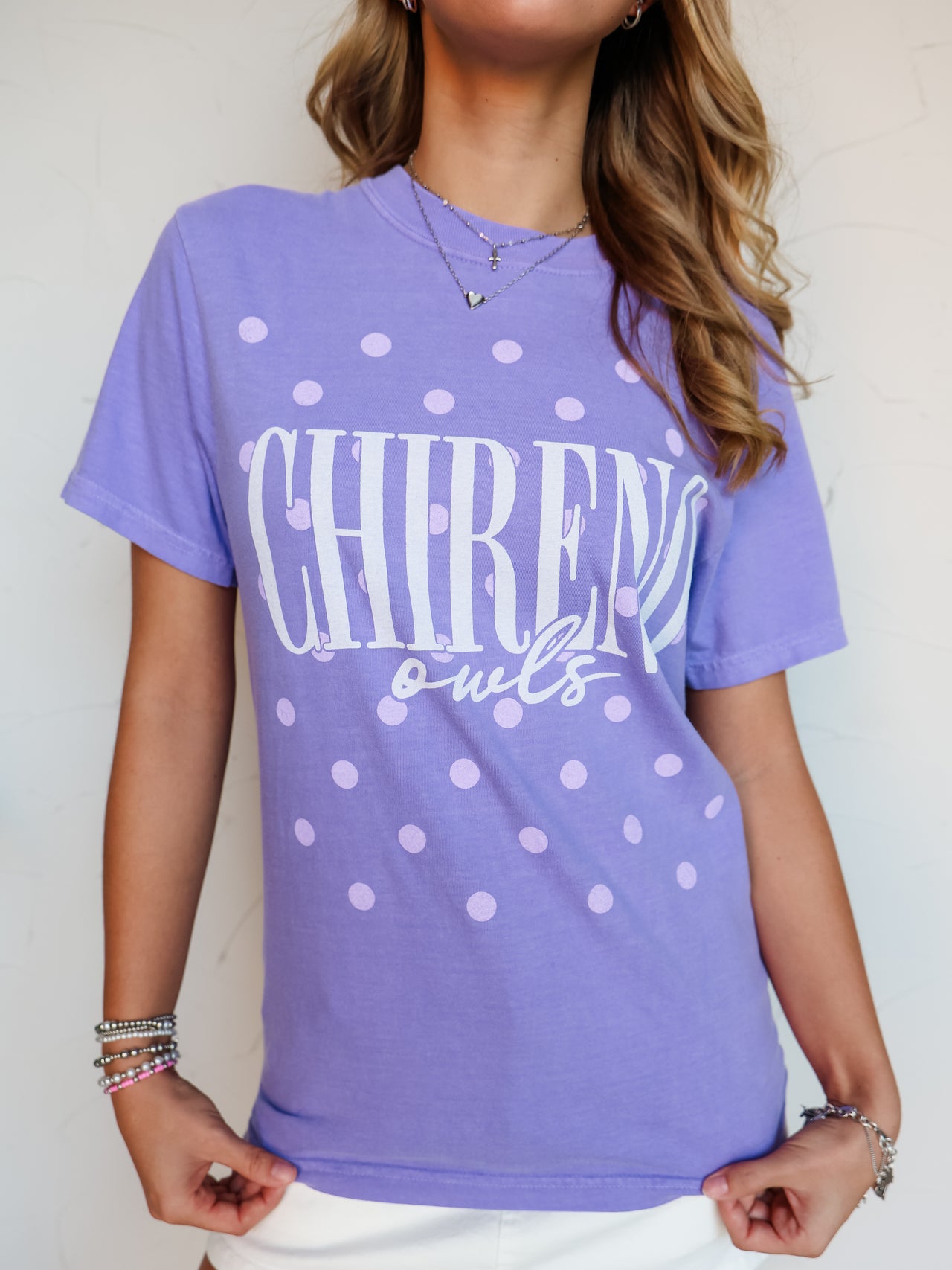 A close-up shot of a person wearing a purple crewneck t-shirt with an all-over lavender polka dot pattern. The shirt features the word "CHIRENO" printed in tall white block letters with "owls" written underneath in a white cursive script font. The person is accessorized with silver necklaces and beaded bracelets, pairing the shirt with white bottoms.