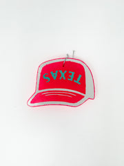 A handmade car air freshener, or freshie, in the shape of a trucker hat, shown against a solid white background. The hat shape is a vibrant neon pink with a light gray border and features the word "TEXAS" embossed across the front in turquoise block letters. A small metal hanging wire is attached at the top.