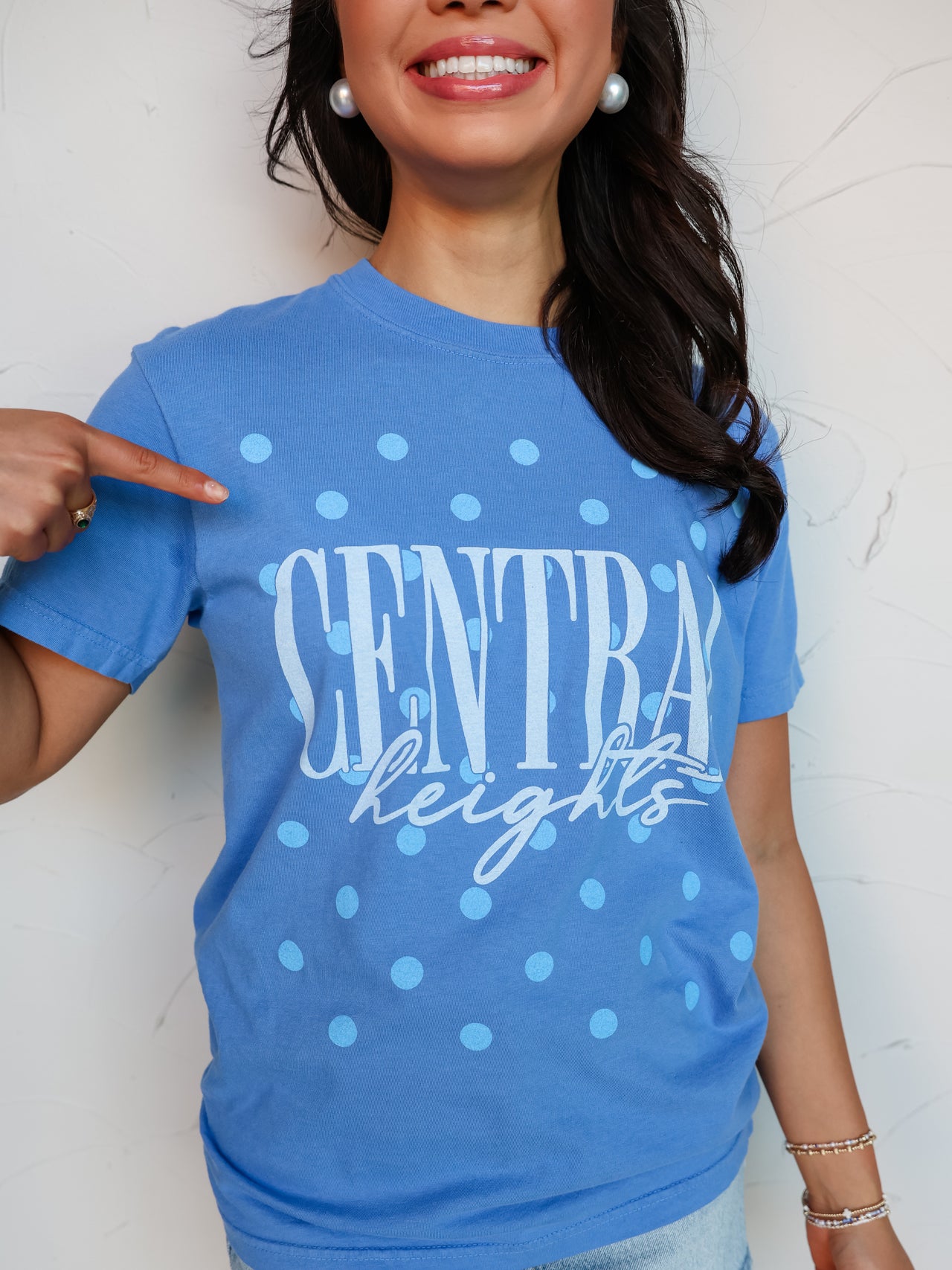 A close-up shot of a smiling person wearing a blue crewneck t-shirt with an all-over light blue polka dot pattern. The shirt features the word "CENTRAL" printed in tall light blue block letters with "heights" written underneath in a white cursive script font. The person is pointing to the graphic on the shirt and accessorized with large white pearl earrings and beaded bracelets.