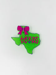 A handmade car air freshener, or freshie, in the shape of the state of Texas, shown against a plain white background. The state cut-out is neon lime green with purple script lettering that reads "Texas" with a small purple heart. A bright magenta bow details the top panhandle section, and a small metal hanging wire is attached.